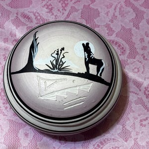 May include: A round, lidded ceramic container with a painted desert scene. The lid features a black and white wolf howling at the moon, with a tree and plants. The container has black and white striped edges.