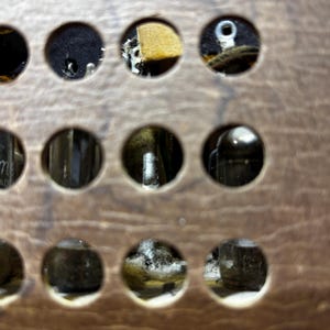 May include: Close-up of a vintage electronic component with multiple circular openings. The openings reveal internal parts, including tubes and wiring, set in a wooden panel. The panel has a warm, brown tone with visible wood grain.