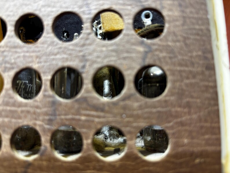 May include: Close-up of a vintage electronic component with multiple circular openings. The openings reveal internal parts, including tubes and wiring, set in a wooden panel. The panel has a warm, brown tone with visible wood grain.