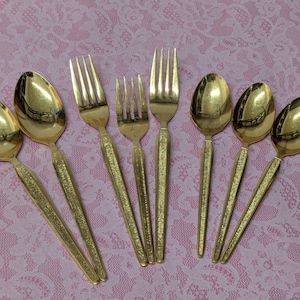 May include: A collection of gold-colored flatware, including spoons and forks, arranged on a pink lace tablecloth. The spoons have oval bowls, and the forks have four tines. The handles have a textured design.