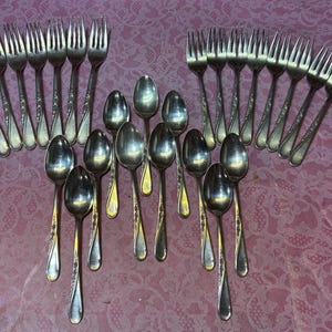 May include: A collection of silver-toned metal flatware consisting of fourteen forks and eleven spoons. Each piece features a slender handle with a subtle embossed floral pattern along the edges. The utensils are arranged in a loose grouping.