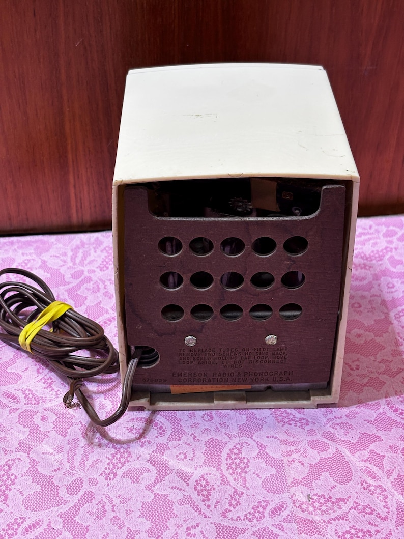 May include: A vintage Emerson Radio & Phonograph, cream-colored with a dark brown speaker panel. The back panel has multiple circular openings and text that reads "Emerson Radio & Phonograph Corporation New York U.S.A."