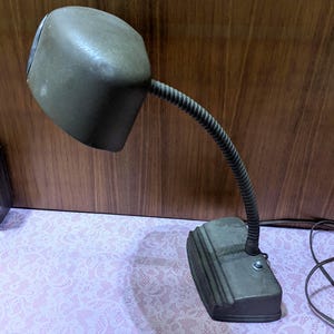 May include: An olive green desk lamp with a flexible, ribbed neck and a rectangular base. The lamp has a rounded, boxy shade and a power cord. The base is textured and has a switch.