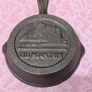 May include: A black cast iron skillet with a handle. The skillet has a raised design of a mountain range and the words "Old Mountain" embossed on the bottom.