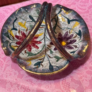 May include: A vintage ceramic serving dish with a handle. The dish has a white background with floral designs in red, purple, yellow, and green. The rim and handle are gold-toned. The dish is on a pink lace tablecloth.