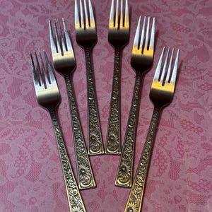 May include: Six silver forks with ornate floral and scrollwork designs on the handles. The tines are polished, and the handles have a gold-toned finish. The forks are arranged on a pink, patterned surface.