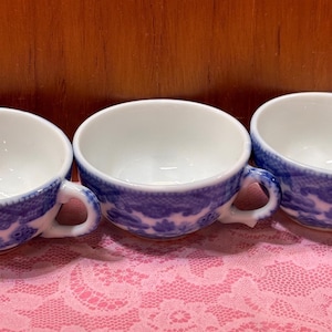 May include: Three white teacups with blue floral patterns and handles are arranged on a pink lace tablecloth. The cups have a classic design, with a blue border and a white interior. The background is a wooden surface.