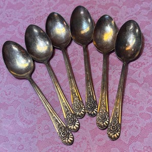 May include: Six antique silver spoons arranged on a pink lace surface. The spoons have ornate floral designs on the handles and oval-shaped bowls. The spoons are made of a shiny, reflective metal.