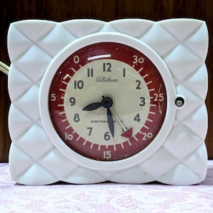 May include: A vintage white Telechron clock with a red and cream-colored face. The clock has a square quilted design around the face and black hands. The clock face displays the numbers 1 through 12 and the brand name Telechron.