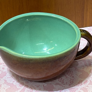 May include: A ceramic gravy boat with a rounded shape and a handle. The interior is a bright teal color, while the exterior transitions from brown to a darker shade. The handle is a dark brown. This kitchenware item is likely used for serving sauces.