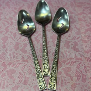 May include: Three silver serving spoons with floral and leaf designs on the handles. Each spoon has a slotted bowl. The spoons are arranged on a pink, patterned surface.