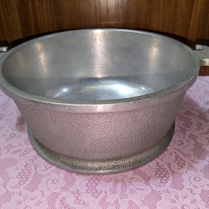May include: A vintage aluminum bowl with a textured exterior and two handles. The bowl has a wide, shallow design and sits on a circular base. The interior is smooth and reflective.