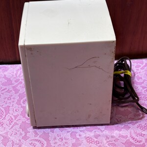 May include: A vintage off-white rectangular speaker with a brown power cord. The speaker has some visible wear and tear, including a crack on the side. The speaker is sitting on a pink floral patterned surface.
