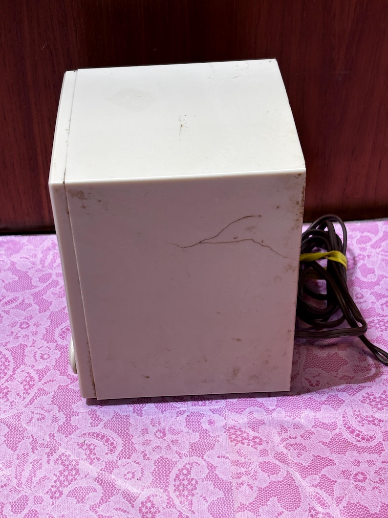 May include: A vintage off-white rectangular speaker with a brown power cord. The speaker has some visible wear and tear, including a crack on the side. The speaker is sitting on a pink floral patterned surface.