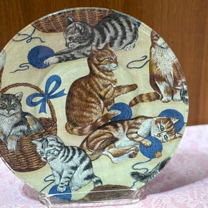 May include: A clear glass plate featuring a whimsical design of various cats. The cats are depicted in different poses, interacting with blue yarn balls and baskets. The plate has a light yellow background and is supported by a clear stand.