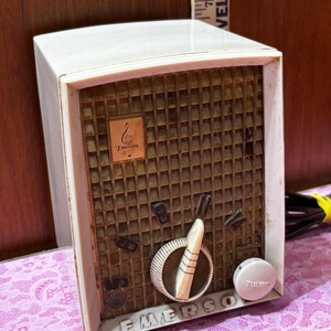 May include: Vintage Emerson radio with a cream-colored plastic casing and a brown speaker grill. The radio features a tuning dial, volume control, and the Emerson logo. The word "EMERSON" is printed on the front.