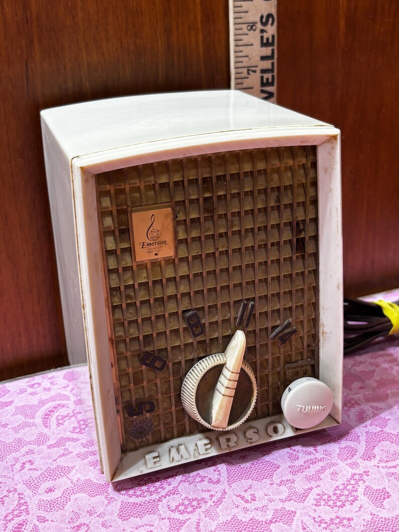May include: Vintage Emerson radio with a cream-colored plastic casing and a brown speaker grill. The radio features a tuning dial, volume control, and the Emerson logo. The word "EMERSON" is printed on the front.