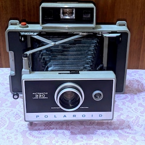 May include: Vintage Polaroid Automatic 330 Land Camera. The camera has a black and silver body with a prominent lens and viewfinder. The words "AUTOMATIC 330 LAND CAMERA" and "POLAROID" are printed on the front. The camera is open, revealing the bellows.