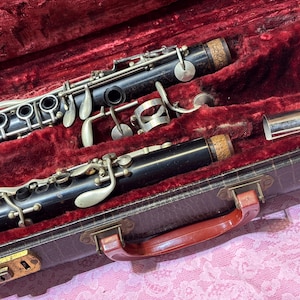 May include: A black clarinet with silver keys and cork accents rests inside a vintage brown case with a red velvet interior. The case has a red handle and brass hardware. The clarinet is partially disassembled.