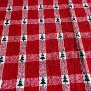 May include: A woven fabric featuring a red and white checkered plaid pattern. Small, dark green pine tree icons are evenly spaced within the white vertical and horizontal stripes, creating a repeating grid design across the entire textile surface.