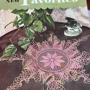 May include: A vintage book cover titled "Old and New Favorites" with a pink crocheted doily and a small ceramic bird figurine. The doily has a floral center and a scalloped edge. A small potted plant is also visible.