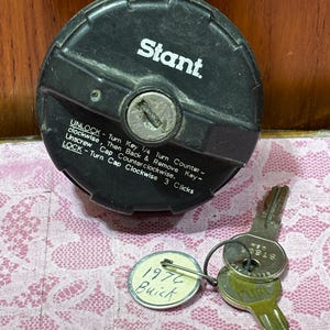 May include: A black circular Stant locking fuel cap with a central keyhole and printed instructions. The text reads: UNLOCK - Turn Key 1/4 Turn Counter-clockwise, Then Back & Remove Key - Unscrew Cap Counterclockwise. LOCK - Turn Cap Clockwise 3 Clicks.