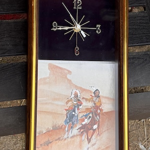 May include: Vintage rectangular wall clock with a gold-colored frame. The top half features a black background with gold-colored clock hands and numbers. The bottom half displays a painting of Native Americans on horseback in a desert landscape.