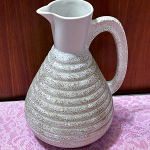 May include: A ceramic pitcher with a light pink top and a textured, beige body. The body features horizontal, ridged detailing. The handle is thick and textured, and the pitcher has a spout for pouring. The pitcher is likely for serving beverages.