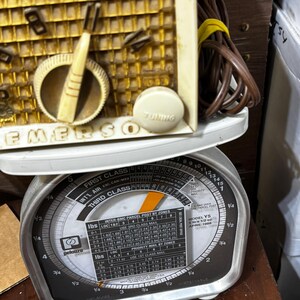 May include: Vintage Emerson radio with a cream-colored face and tuning dial, resting on a Pelouze postal scale. The scale displays weight measurements in pounds and ounces, with a dial for calculating postage.