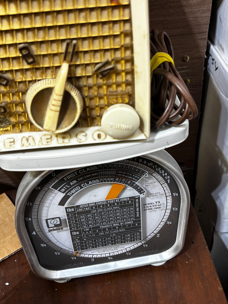 May include: Vintage Emerson radio with a cream-colored face and tuning dial, resting on a Pelouze postal scale. The scale displays weight measurements in pounds and ounces, with a dial for calculating postage.
