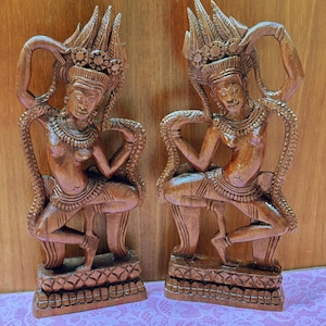 May include: Two wooden sculptures of dancing figures. The figures are carved in a detailed style, with ornate headdresses and jewelry. The sculptures are a warm brown color and are set against a wooden background.
