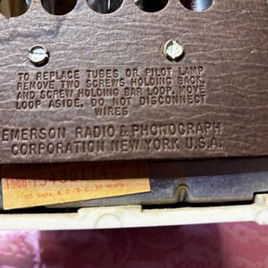 May include: Close-up of a vintage Emerson Radio & Phonograph Corporation component. The brown panel features text instructions and the company name. A small sticker displays model and voltage information. The background is a cream-colored frame.