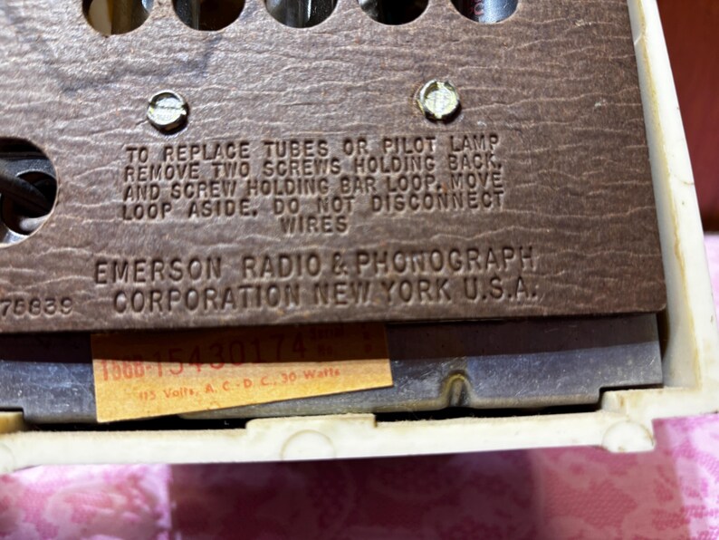 May include: Close-up of a vintage Emerson Radio & Phonograph Corporation component. The brown panel features text instructions and the company name. A small sticker displays model and voltage information. The background is a cream-colored frame.