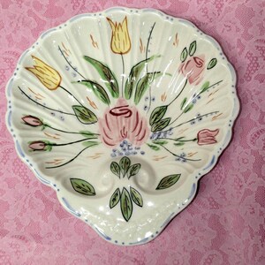 May include: A white, shell-shaped ceramic dish with a scalloped edge and a light blue border. The dish is decorated with a floral design featuring pink and yellow tulips, green leaves, and small blue accents. The dish has two small indentations.