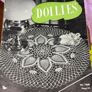 May include: A vintage book cover titled "DOLLIES" with a green banner. The cover features a white crocheted doily and a lamp. The text reads "A Collection of Fascinating New Designs".