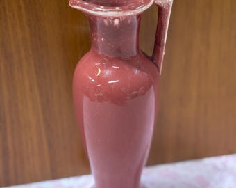 May include: A maroon-colored ceramic pitcher with a handle. The pitcher has a narrow neck and a flared spout. The handle is rectangular and attached to the side of the pitcher. The pitcher is approximately 7 inches tall.