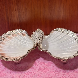 May include: A decorative seashell dish with two white shell-shaped bowls, each with a silver-colored trim. A smaller shell is in the center. The dish is supported by three silver-colored ball feet. The dish is on a pink lace surface.