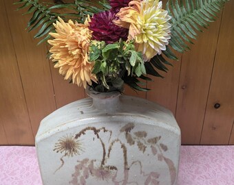 May include: A bouquet of yellow, white, and burgundy dahlias with green ferns in a white ceramic vase with a brown floral design.