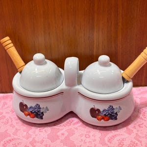 May include: White ceramic double condiment server with lids and wooden handled spoons. The server has a handle and is decorated with a fruit design of apples, grapes, and strawberries. A red stripe accents the rim.