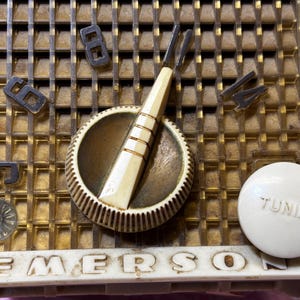 May include: Close-up of a vintage Emerson radio. The radio features a gold-colored grill with the numbers 5, 6, 8, and 14. A white tuning knob is visible, along with a cream-colored dial and a tuning needle. The word "EMERSON" is printed on the bottom.
