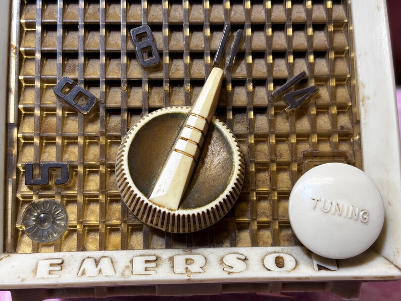 May include: Close-up of a vintage Emerson radio. The radio features a gold-colored grill with the numbers 5, 6, 8, and 14. A white tuning knob is visible, along with a cream-colored dial and a tuning needle. The word "EMERSON" is printed on the bottom.