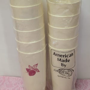 May include: Two stacks of off-white paper cups, each wrapped in clear plastic. One stack features a magenta strawberry graphic. The other stack has a label that reads "American Made By NASCO inc. Springfield, Tn."