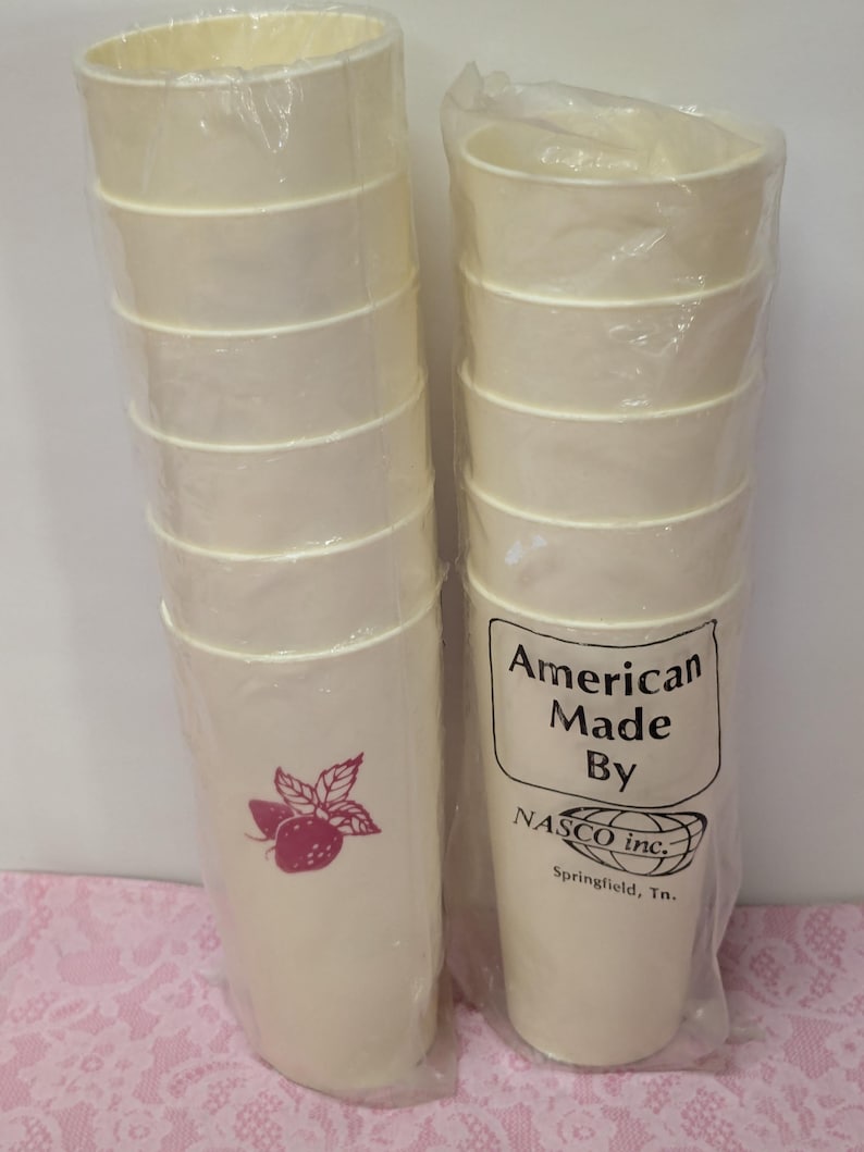 May include: Two stacks of off-white paper cups, each wrapped in clear plastic. One stack features a magenta strawberry graphic. The other stack has a label that reads "American Made By NASCO inc. Springfield, Tn."