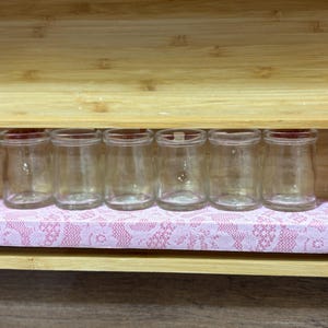 May include: Six small, clear glass jars with red lids are arranged on a pink lace-patterned shelf inside a wooden box. The jars are cylindrical with a slightly wider base. The wooden box has a natural wood finish.