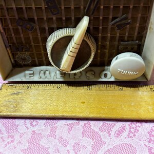 May include: Close-up of a vintage Emerson radio. The radio has a cream-colored casing with a brown speaker grill. The word "EMERSON" is printed on the front. A tuning knob and dial are visible. A wooden ruler is in the foreground.