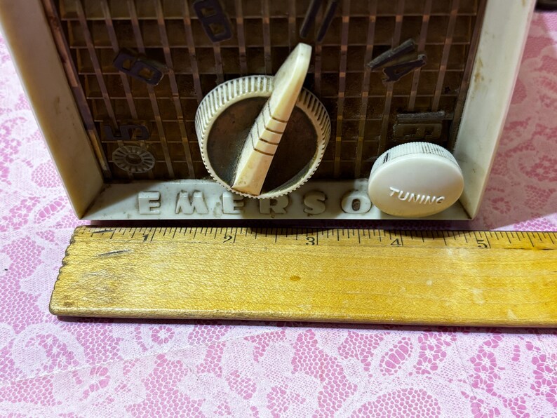 May include: Close-up of a vintage Emerson radio. The radio has a cream-colored casing with a brown speaker grill. The word "EMERSON" is printed on the front. A tuning knob and dial are visible. A wooden ruler is in the foreground.