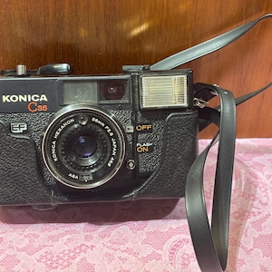 May include: Black Konica C35 camera with a leather strap. The camera has a silver lens and a built-in flash. The text "KONICA C35" is printed on the top of the camera. The lens reads "KONICA HEXANON 38mm F2.8 JAPAN 46".
