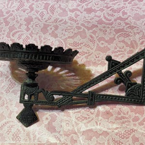 May include: An antique black metal wall sconce with a decorative, round, scalloped tray and ornate bracket. The sconce has a diamond-shaped base and intricate scrollwork details. The tray is approximately 8 inches in diameter.