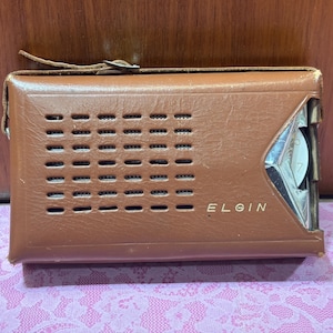 May include: A vintage brown leather-cased radio with the brand name "ELGIN" printed on the front. The radio has a speaker grill with multiple rectangular openings and a silver-colored dial. A leather strap is attached to the top.