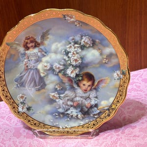 May include: Decorative plate featuring two cherubic figures with wings amidst clouds and flowers. The plate has a scalloped edge with a gold rim and a gold floral border. The artwork is in soft pastel colors.